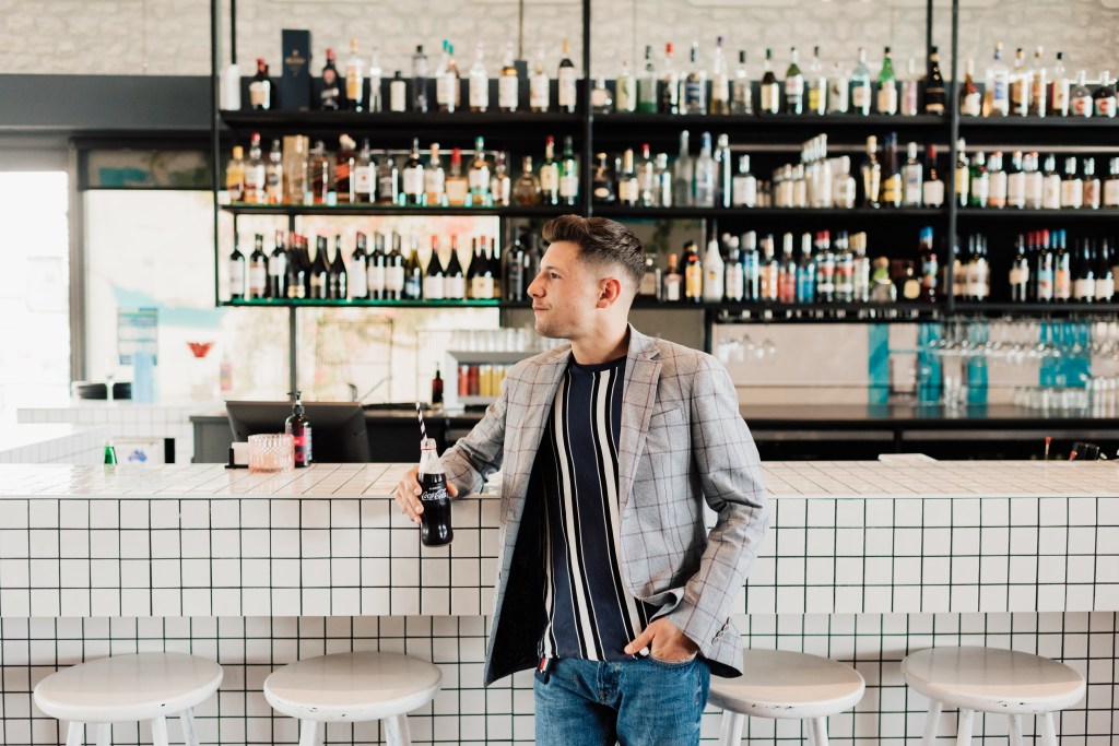 Man stands at the bar at Ellenika Ocean Grill with a glass bottle of Coca-Cola in hand.